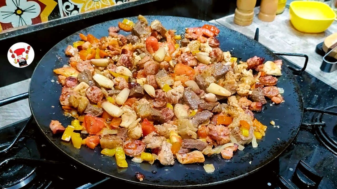 Panela grande com entrevero, mostrando pedaços de carne bovina, frango, linguiça, batata, cenoura e pimentões coloridos.