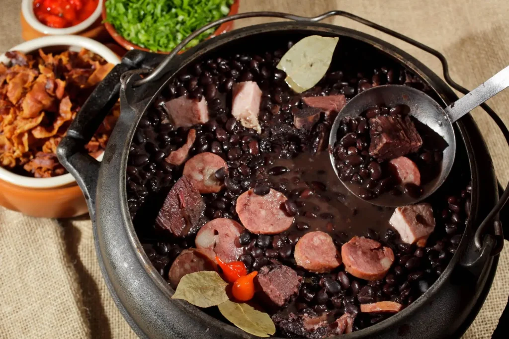 Panela grande de feijoada com carnes variadas, servida com arroz, couve, farofa e laranja.