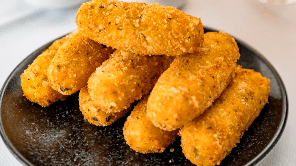 Foto apetitosa de um prato com diversos bolinhos de milho verde fritos e dourados, prontos para serem consumidos, com milho verde espalhado ao redor.