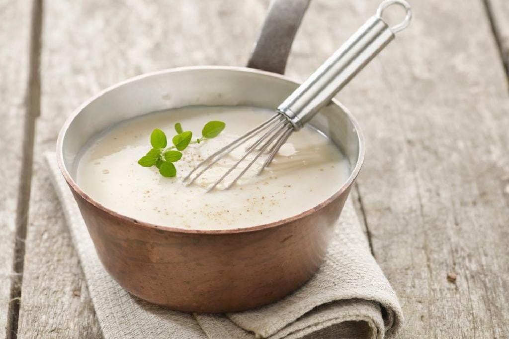 Panela grande com molho branco cremoso, sendo mexido com um fouet.