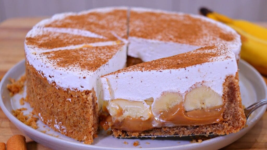 Fatia de banoffee em um prato, mostrando as camadas de biscoito, doce de leite, banana e chantilly, polvilhada com cacau em pó.