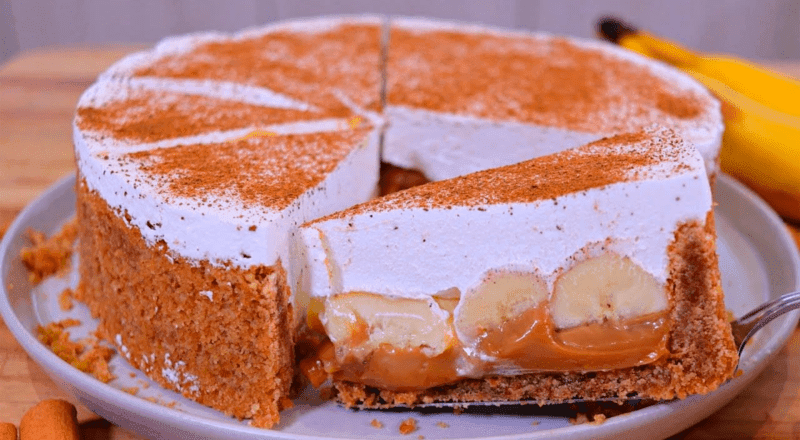 Fatia de banoffee em um prato, mostrando as camadas de biscoito, doce de leite, banana e chantilly, polvilhada com cacau em pó.