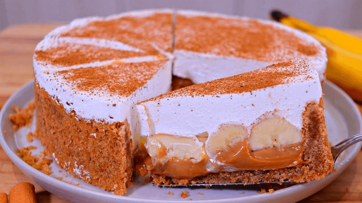 Fatia de banoffee em um prato, mostrando as camadas de biscoito, doce de leite, banana e chantilly, polvilhada com cacau em pó.