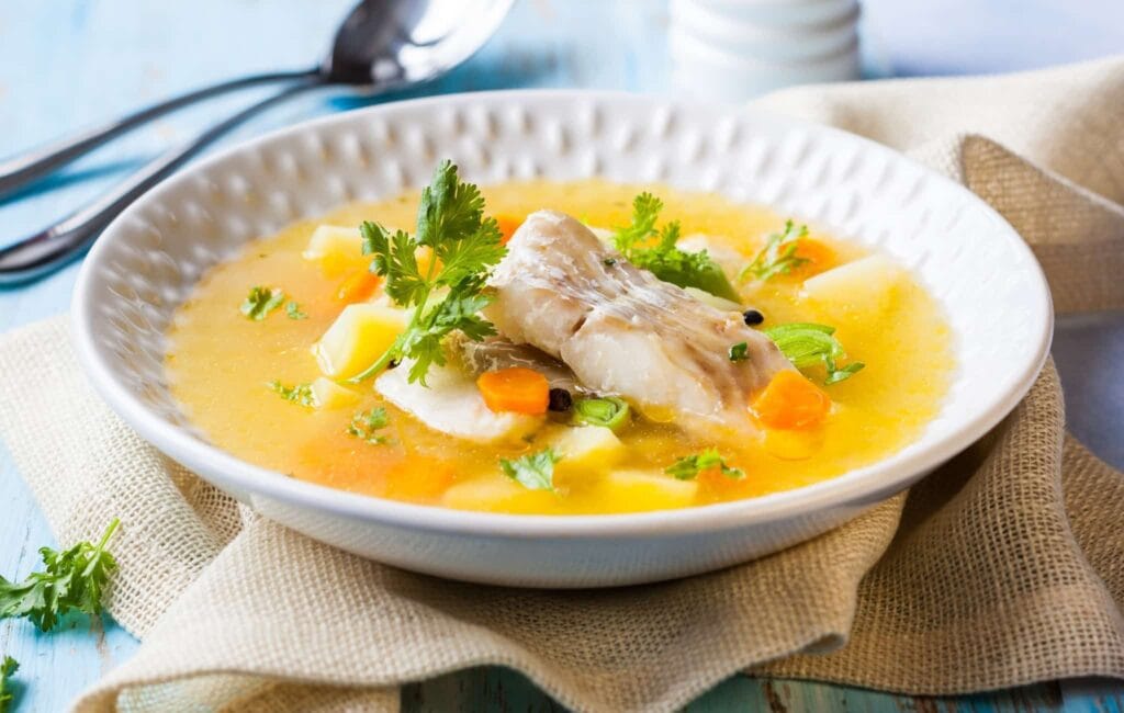 Tigela de sopa de peixe tailandesa fumegante, com pedaços de peixe branco, vegetais coloridos e cebolinha picada, em um caldo aromático.