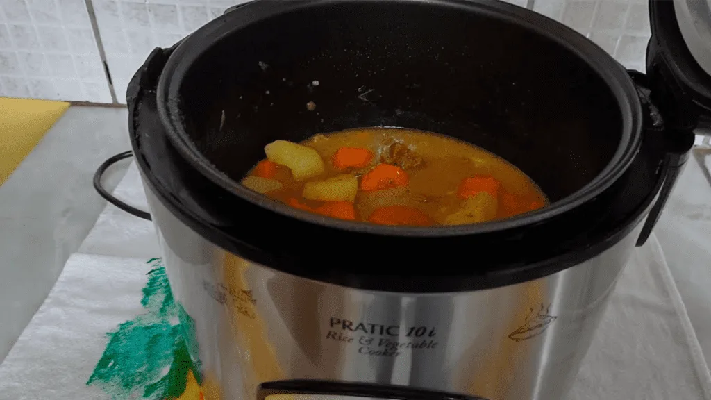 Uma panela elétrica de arroz aberta, mostrando pedaços de carne cozida e suculenta com legumes, representando o resultado final da receita e a praticidade do método.