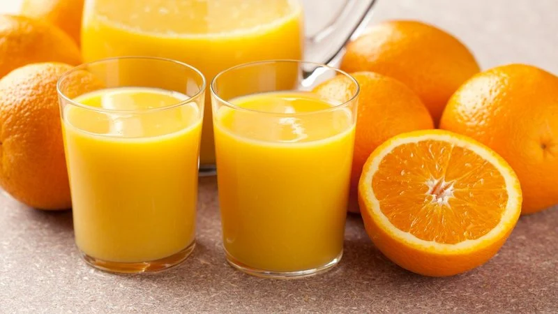 Um copo de suco de laranja fresco e vibrante, com laranjas ao fundo, representando a bebida refrescante e o tema do artigo.