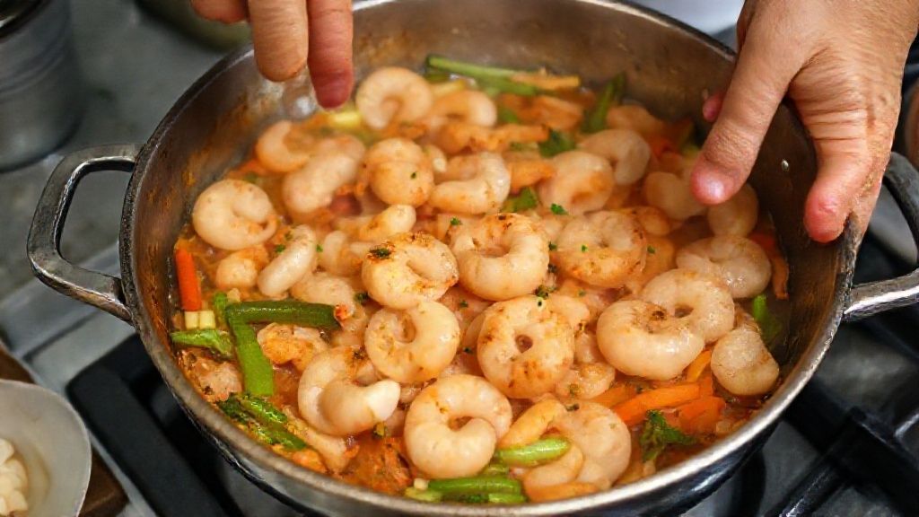 Mãos refogando camarões e verduras em panela durante o preparo do bobó.