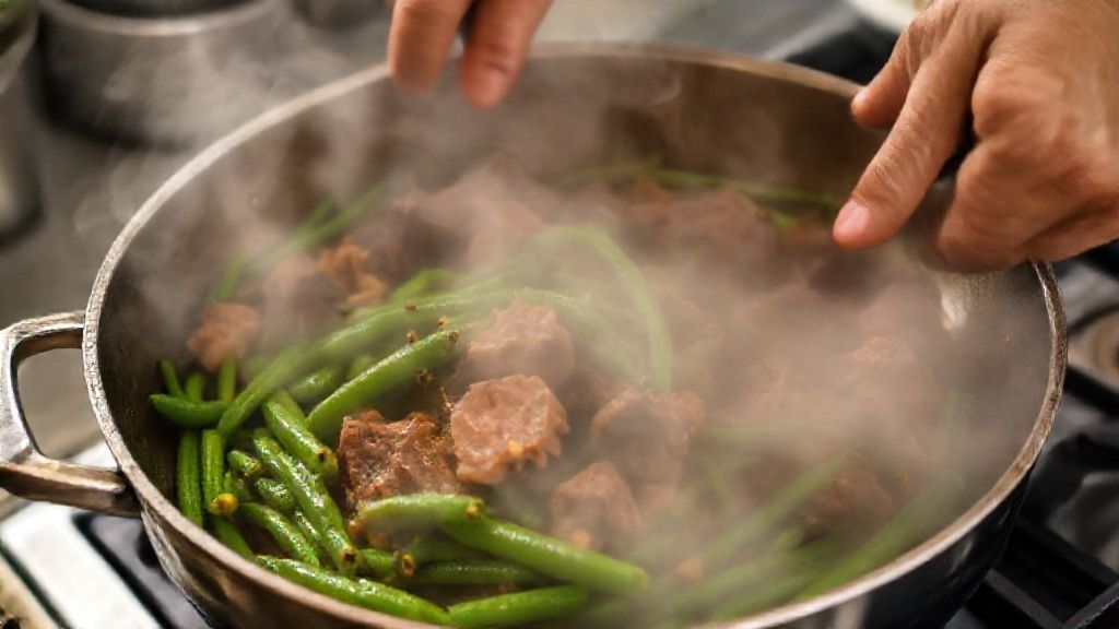 Mãos adicionando vagens à panela com carne refogada, mostrando vapor e brilho dos ingredientes cozinhando juntos