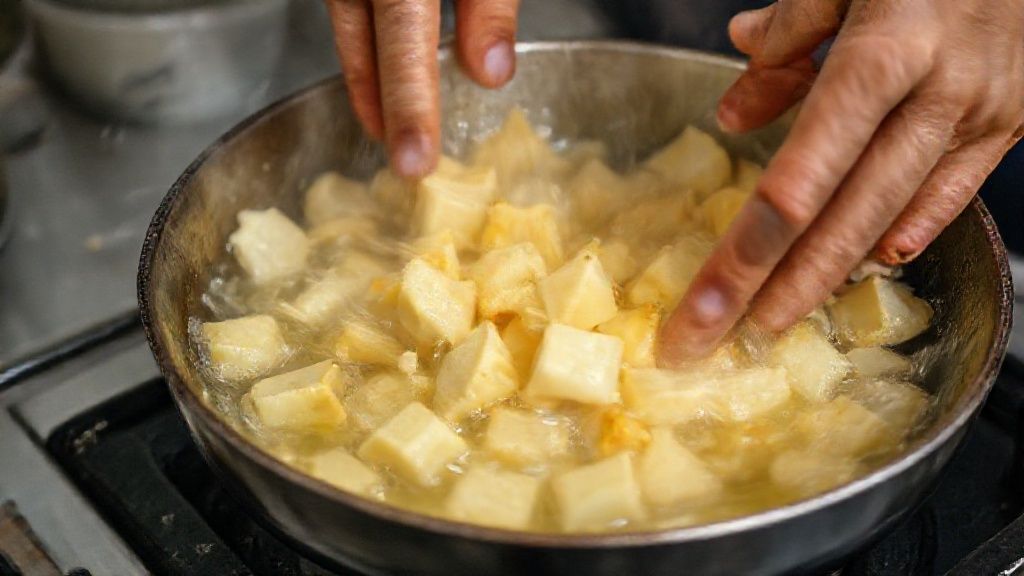 Mãos mergulhando mandiocas pré-cozidas em óleo quente, mostrando a etapa de fritura dourada.