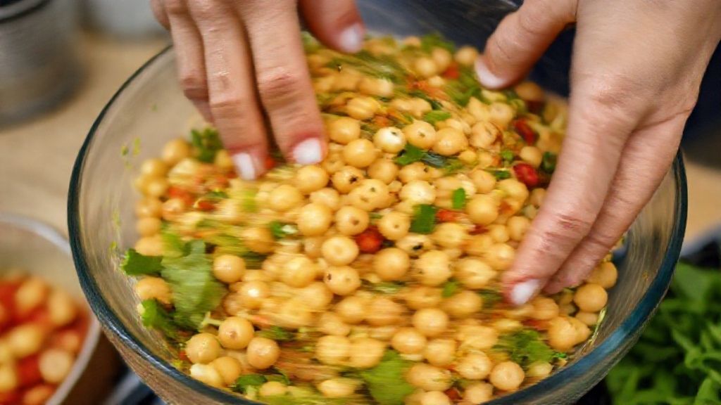 Mãos misturando o grão de bico cozido com legumes em uma tigela grande durante o preparo