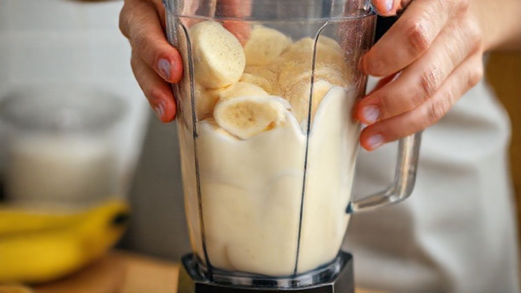 Mãos adicionando bananas e leite condensado ao liquidificador durante o preparo da sobremesa