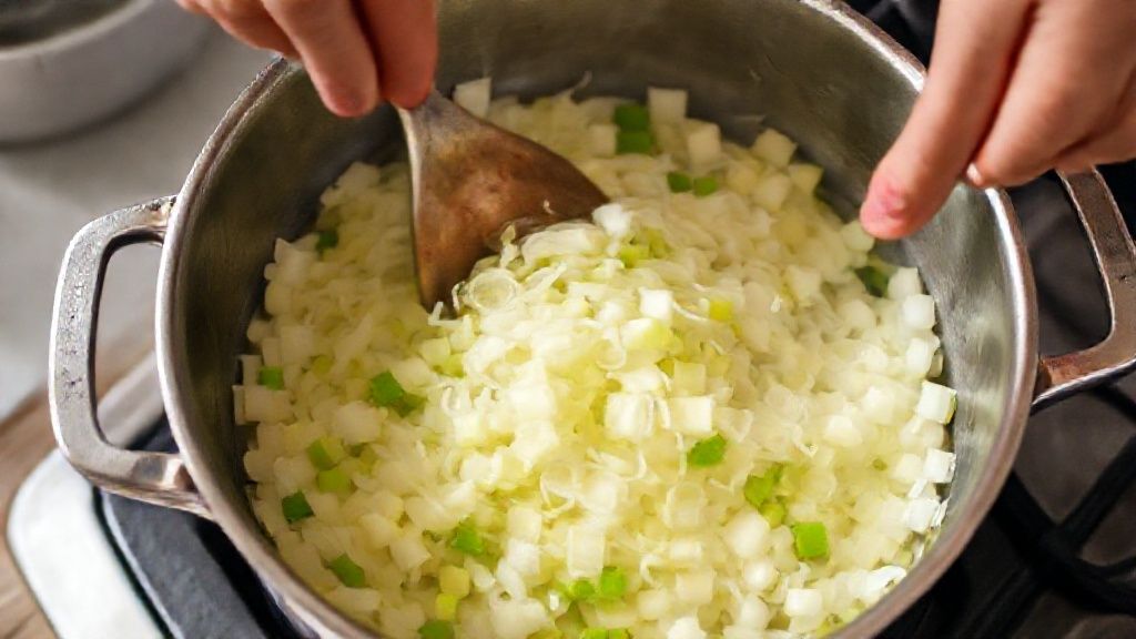 Mãos refogando alho-poró e cebola em panela de fundo grosso, passo essencial para realçar o sabor da sopa.