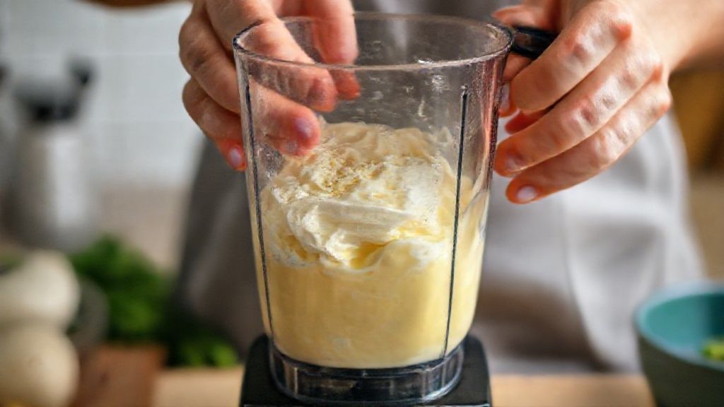 Mãos batendo os ingredientes do bolo pudim no liquidificador em cozinha iluminada