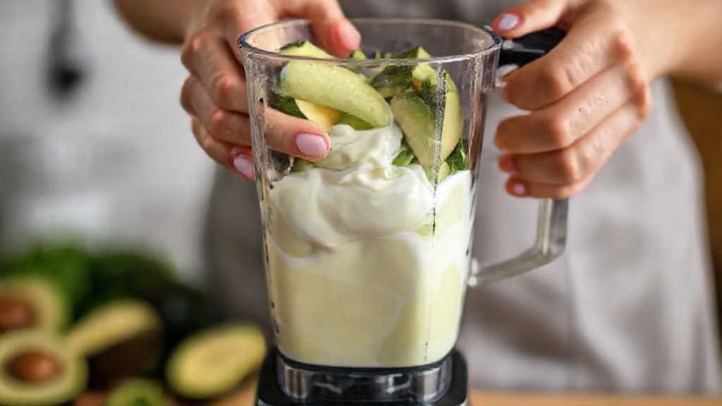 Mãos colocando pedaços de abacate no liquidificador e misturando os ingredientes.