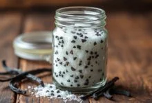Um pote de vidro transparente cheio de açúcar cristal perfumado, com sementes de baunilha escuras visíveis e vagens inteiras repousando ao lado, sobre uma superfície rústica de madeira.