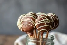 Alfajores no palito com cobertura de chocolate ao leite brilhante e decoração de chocolate branco, prontos para servir.