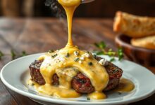 Fatia de bife grelhado coberta por um molho 4 queijos dourado e elástico, com fios de muçarela e pedaços de gorgonzola visíveis.