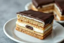 Fatia de bolo de biscoito com chocolate em primeiro plano, mostrando camadas uniformes de biscoito maisena e creme firme, coberto com uma camada espessa e brilhante de chocolate meio amargo.