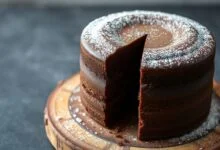 Um bolo de chocolate alto, com cobertura lisa e cor marrom profundo, fatiado para revelar a textura fofinha e úmida do interior.