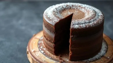 Um bolo de chocolate alto, com cobertura lisa e cor marrom profundo, fatiado para revelar a textura fofinha e úmida do interior.