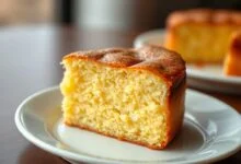 Fatia generosa de bolo de mandioca com casquinha dourada, miolo úmido e cremoso, servida em um prato de porcelana branca.