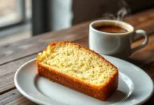 Fatia generosa de bolo de manteiga dourado, com textura fofa e macia, servida em um prato de porcelana branca ao lado de uma xícara de café.