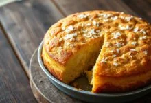 Bolo de milho com coco inteiro em forma redonda, dourado e com textura úmida, fatiado e servido em prato branco.