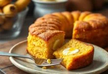 Um bolo de milho dourado, com uma fatia generosa servida ao lado, revelando o interior úmido e os cremosos bolsões de cream cheese.