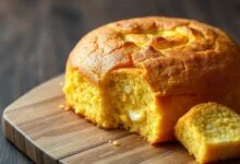 Bolo de milho de liquidificador inteiro, com superfície dourada e textura cremosa visível, fatiado sobre uma tábua de madeira.