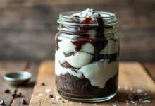 Pote de vidro transparente com camadas generosas de bolo fofo de cacau escuro, beijinho cremoso de coco e uma calda brilhante por cima.