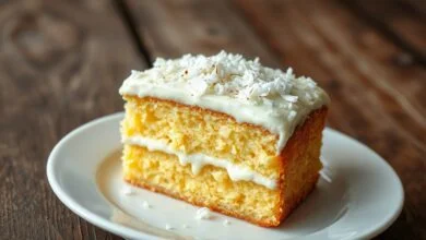 Fatia generosa de bolo gelado de coco com camadas úmidas, calda cremosa visível e raspas de coco fresco, servido em prato branco sobre mesa rústica.