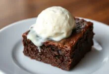 Fatia de brownie fudgy com casquinha crocante, centro úmido e denso, coberta por uma bola generosa de sorvete de baunilha derretendo lentamente, servida em um prato branco.