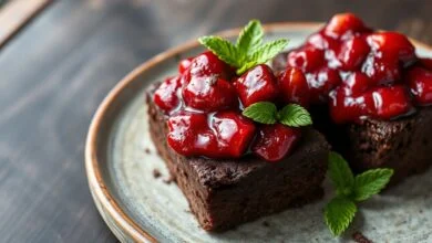 Fatias de brownie de cacau escuro e denso, com textura úmida visível no corte, cobertas por uma calda brilhante e espessa de morangos frescos picados, dispostas sobre um prato de cerâmica rústica.