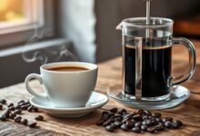 Xícara de café encorpado servido ao lado da prensa francesa de vidro, com tons de caramelo e creme na superfície.
