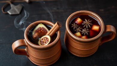 Duas canecas de barro fumegantes, uma com quentão escuro e figos, outra com vinho quente e maçãs, sobre superfície rústica.