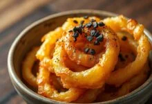 Rodelas de cebola douradas com bordas crocantes e centro translúcido, finalizadas com pimenta-do-reino em tigela rústica.