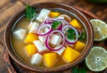 Tigela de ceviche fresco com cubos de peixe branco opaco, manga amarela vibrante e anéis de cebola roxa, servido em uma tigela de barro sobre uma mesa de madeira rústica.