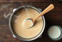Panela com molho cremoso sendo mexido com colher de madeira, com um potinho de creme de leite ao lado