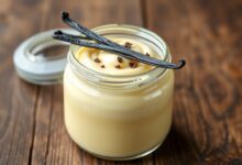 Um pote de vidro transparente repleto de creme de confeiteiro amarelo pálido, liso e brilhante, com uma baunilha inteira ao lado e uma pitada de raspas de baunilha no topo.