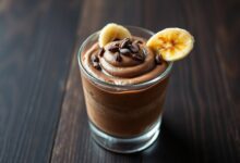 Uma taça de vidro transparente revelando a textura aveludada do creme marrom escuro, finalizada com lascas de cacau e uma rodelas de banana caramelizada na borda.