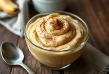 Tigela de vidro com doce de banana cremoso de cor caramelo homogênea, textura espessa e finalizado com um toque de canela em pó.