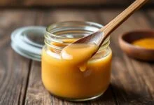 Um pote de vidro aberto, transbordando com doce de leite caseiro de cor caramelo dourado, com textura lisa e cremosa. Uma colher de madeira está mergulhada no doce, mostrando a consistência perfeita.