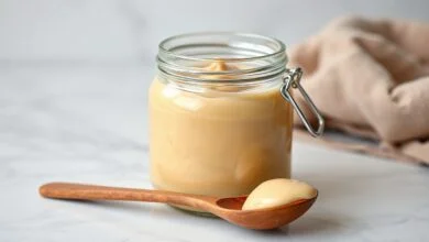 Um pote de vidro transparente e cheio de doce de leite Ninho, de cor bege clara e textura homogênea e cremosa, com uma colher de madeira apoiada ao lado, sobre uma superfície de mármore.