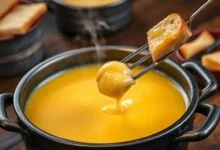 Panela de fondue com queijo derretido, homogêneo e levemente dourado, borbulhando suavemente, pronta para mergulhar pães e legumes.