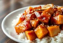 Cubos de frango dourados cobertos por molho agridoce caramelizado e brilhante, com sementes de gergelim, fatias de cebola e alho, servidos ao lado de arroz branco soltinho.
