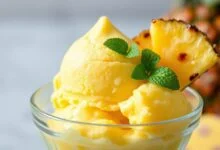 Uma taça de vidro com uma bola generosa de gelado de abacaxi amarelo vibrante e textura aveludada, decorada com um pedaço de abacaxi fresco e folha de hortelã.
