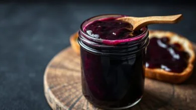 Pote de vidro com geleia de jabuticaba de cor roxa intensa e brilhante, com uma fatia de pão ao fundo.