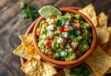 Tigela de guacamole cremoso com pedaços de abacate, tomate e coentro fresco, acompanhada de tortilhas chips crocantes.