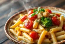 Uma tigela fumegante de macarrão penne coberto por um molho cremoso e encorpado, com tomates cereja levemente rachados, brócolis vibrante e pedaços de alho assado dourado.