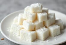 Cubos de maria-mole branca e fofa, cobertos com flocos de coco ralado, dispostos sobre um prato de porcelana.